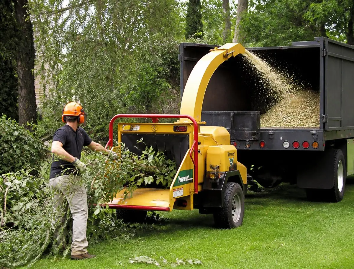 Ouvrier casqué engageant des branches dans un broyeur ; les copeaux sont projetés dans la benne d’un camion